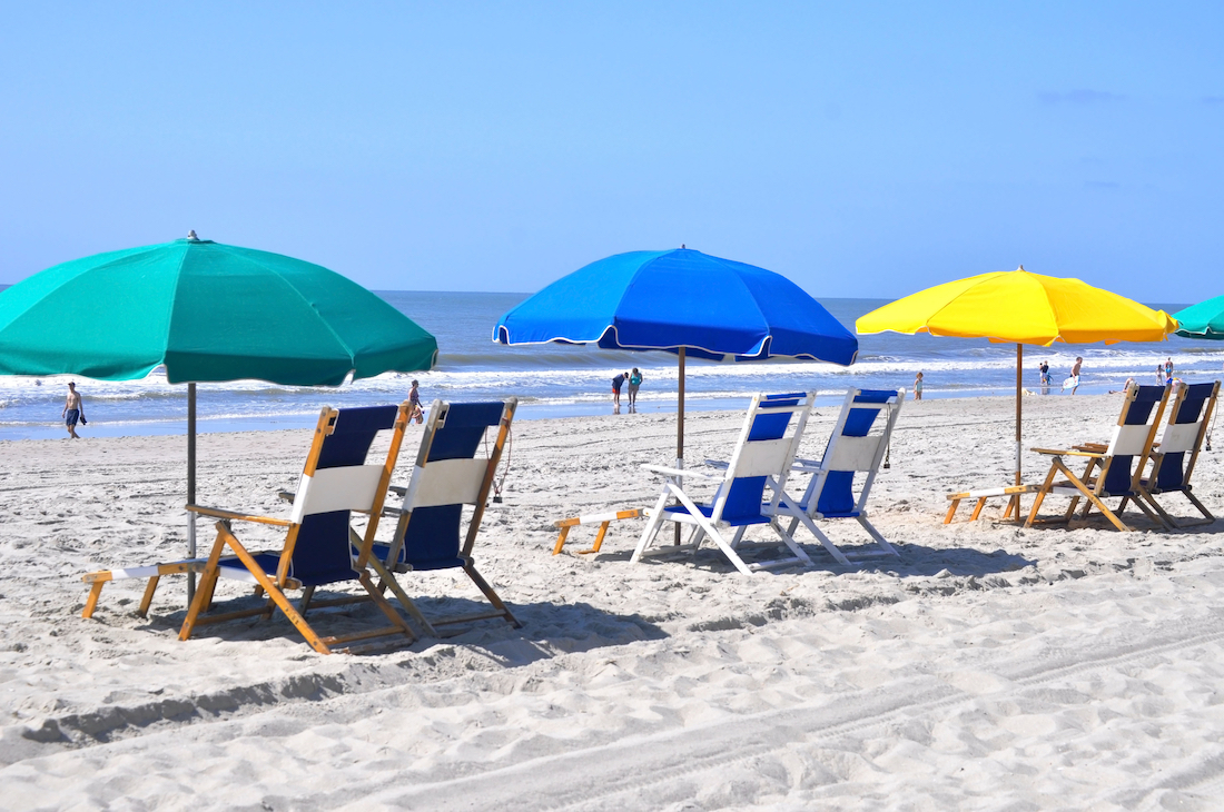 Green, blue, and yellow beach umbrellas cover pairs of beach chairs all resting in the sand