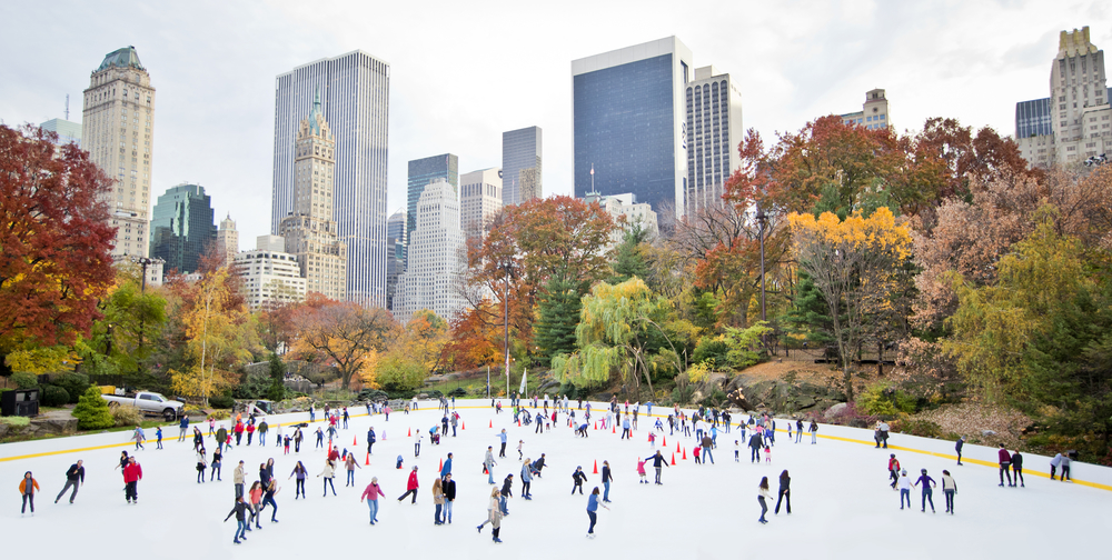 Crowds ice skating at Wollman Rink in Central Park with building peering through trees in background