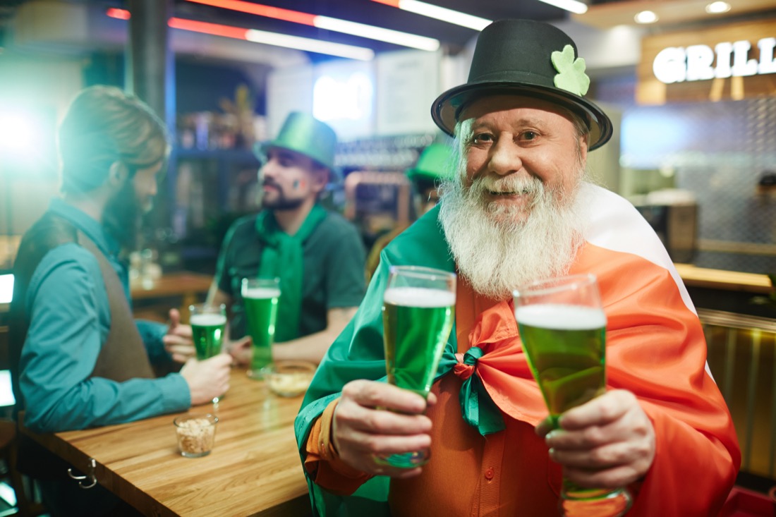 Senior man in hat holding beer with Irish flag on shoulders celebrating St Patrick's Day