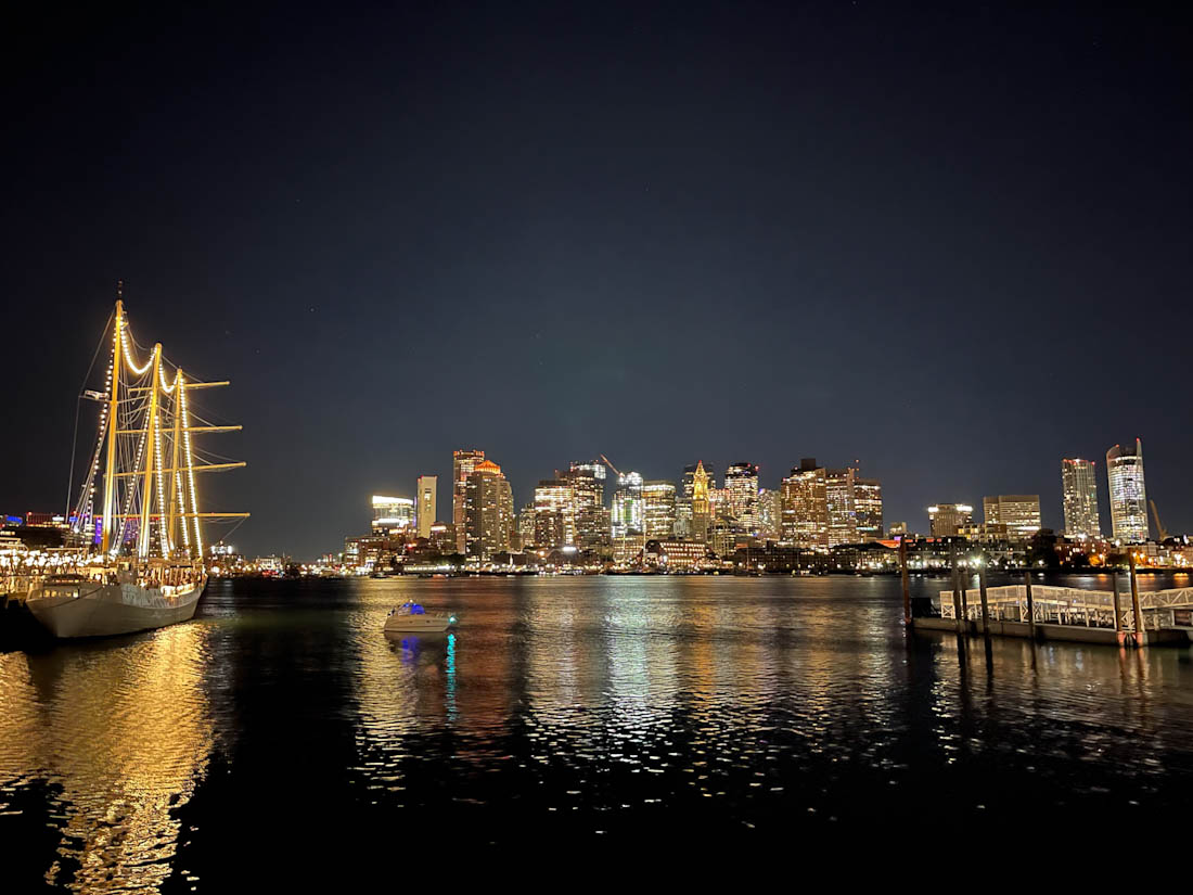 A boat lit up in the water at night with the city skyline beyond, seen from East Boston