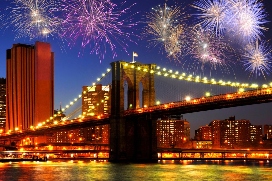 NYC skyline at night with colorful fireworks over Brooklyn bridge.