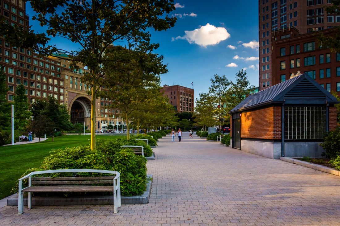 View of the Rose Fitzgerald Kennedy Greenway in Boston with a bench, surrounding buildings, and people jogging.
